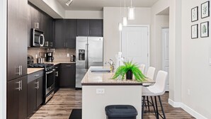 A modern kitchen with dark cabinets, stainless steel appliances, a central island with a sink, and a dining area with white chairs. - Charming Tempe 1BR Designer Furnishings, Patio & Gym (Tempe)