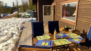 A wooden deck with a table set for dining, surrounded by chairs, and a view of a snowy landscape with parked cars. - Magical west view in Sälen, 8 beds in 3 bedrooms in cozy spacious log cabin (Dalarnas län)