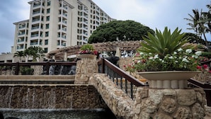 A multi-story hotel with a stone bridge over a water feature, surrounded by lush greenery and a pergola.