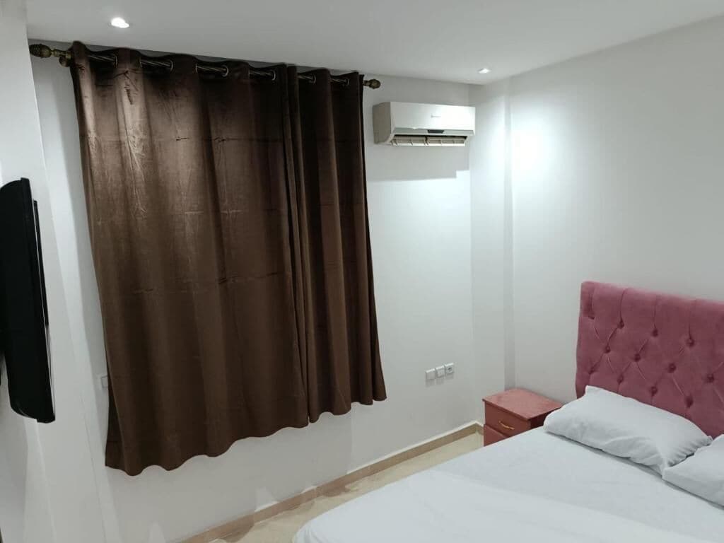 A bedroom with a bed, a nightstand, a red headboard, a brown curtain, and an air conditioning unit.