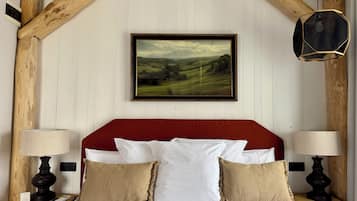 Una cama con sábanas blancas y dos almohadas, cabecera roja, vigas de madera, un cuadro de paisaje enmarco y dos mesitas de noche con lámparas.