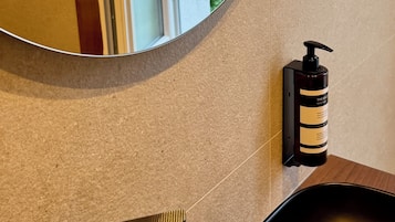 Ein Badezimmer mit einem runden Spiegel, einem schwarzen Stuhl, einer Shampooflasche und Blick auf einen Balkon mit Grünpflanzen und Bergen.