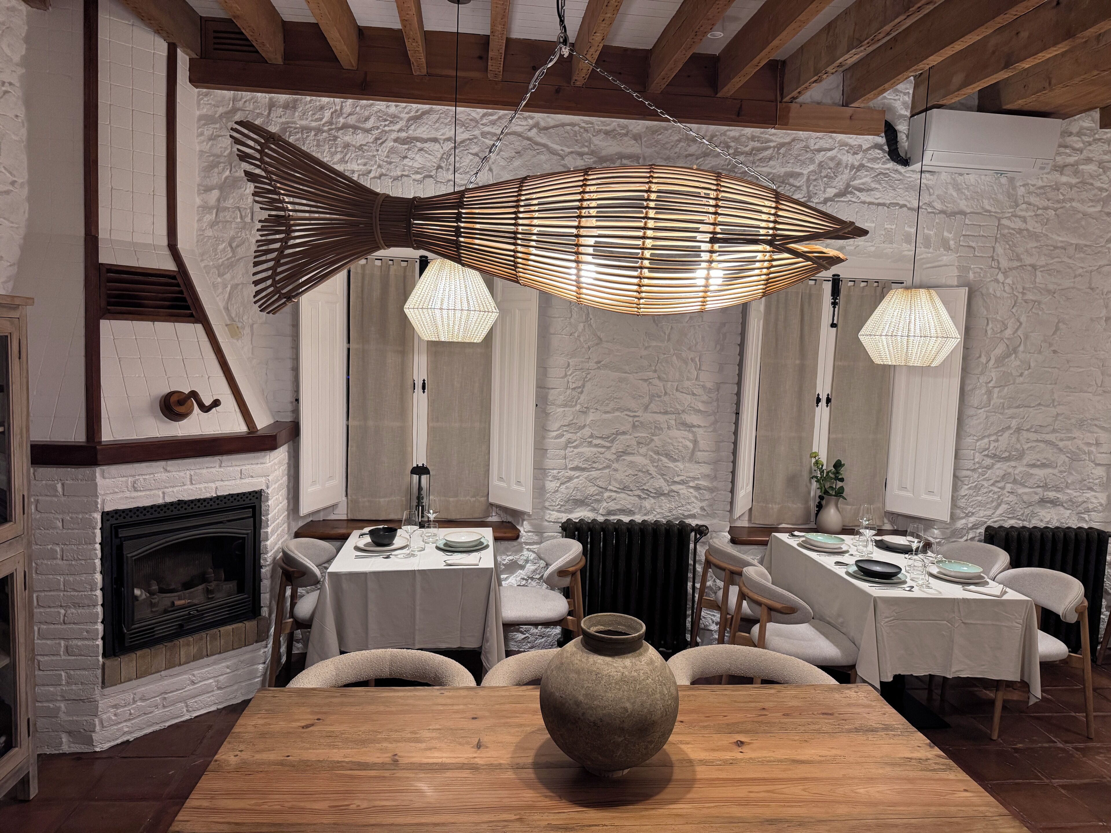A dining area with a rustic fireplace, wooden beams, and a large fish-shaped hanging light fixture.