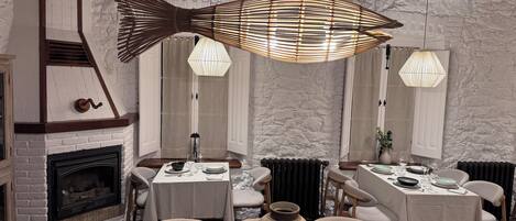 A dining area with a rustic fireplace, wooden beams, and a large fish-shaped hanging light fixture.