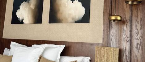 A hotel room with a wooden headboard, a bed with white and beige bedding, two wall-mounted paintings, and a table lamp.