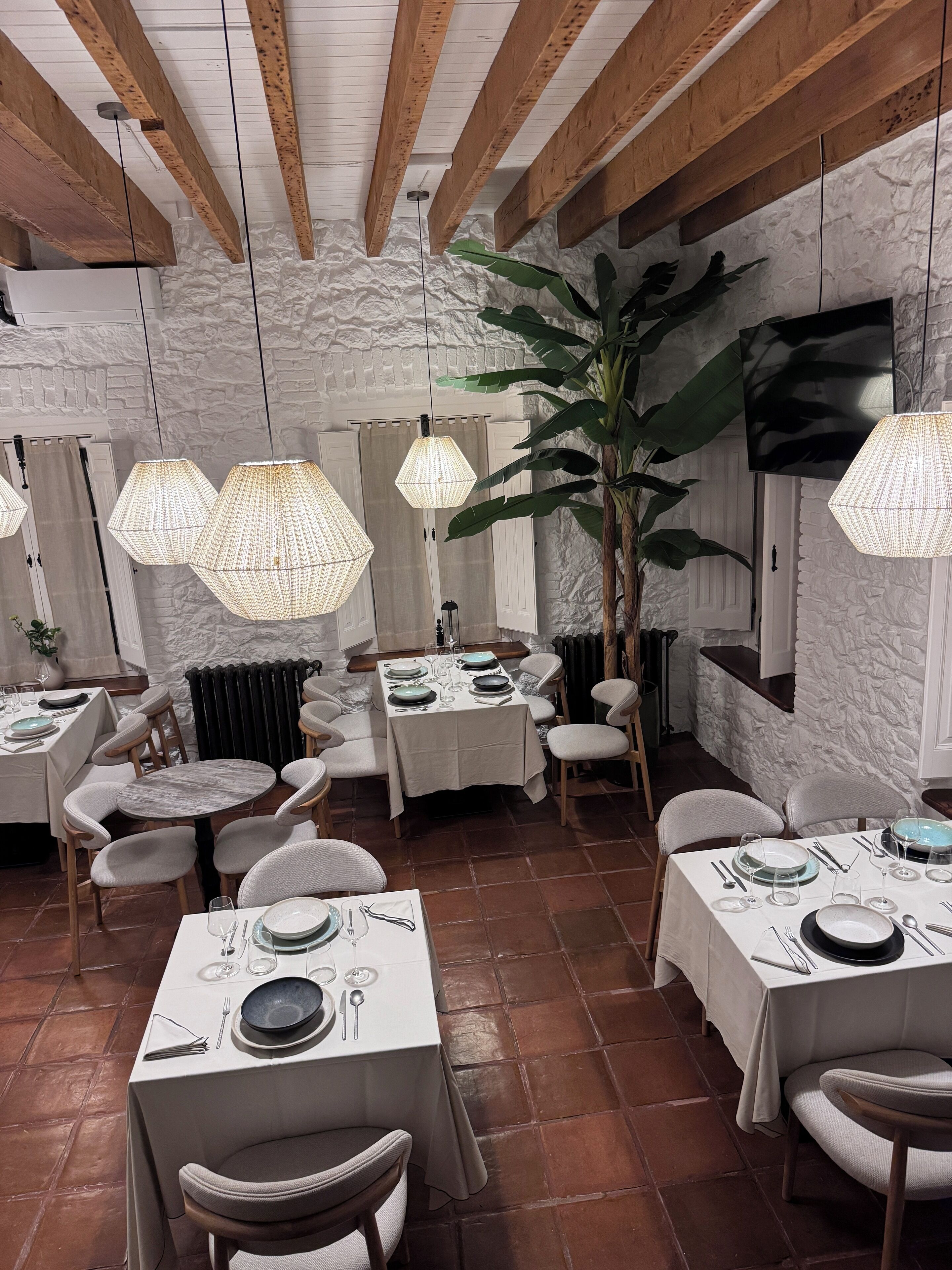 Un restaurante con vigas de madera rústicas, paredes de piedra y lámparas de papel colgantes. Las mesas están preparadas con platos, cubiertos y vasos.