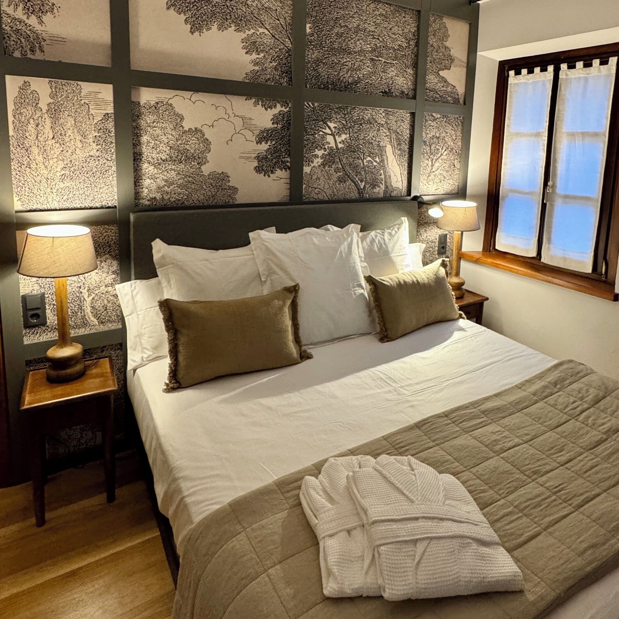 A neatly made bed with white linens and two pillows, a quilted blanket, and a folded robe on the bed. There are two bedside lamps on wooden nightstands.