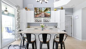 Una cocina moderna con una isla central, armarios blancos y un televisor grande sobre la isla que muestra una escena de playa.
