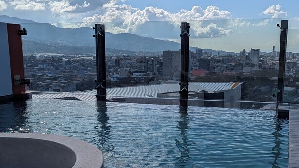 Una piscina infinita con vistas a la ciudad, un área de estar circular y una barandilla de vidrio.