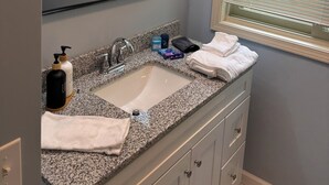 A bathroom with a granite countertop sink, a mirror with lights, a window with blinds, and a towel rack.