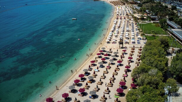 An aerial view of a beach with rows of sun loungers and umbrellas, a clear blue sea, and a cityscape in the background.