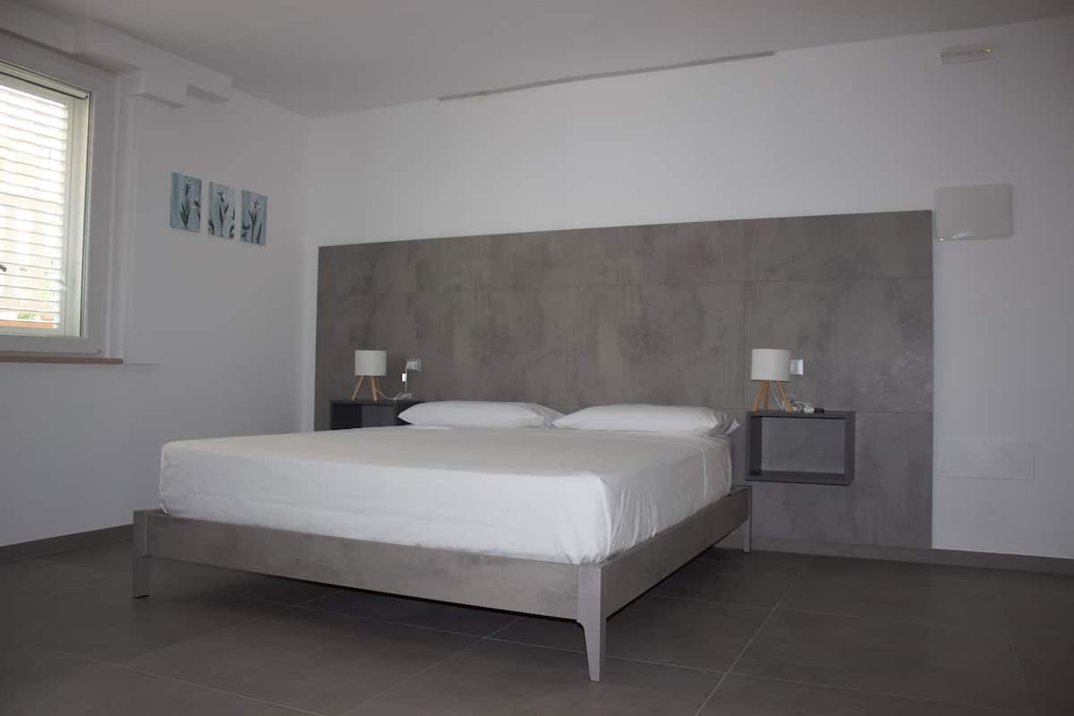 A modern bedroom with a grey headboard, a large bed with white linens, two bedside tables with lamps, and a window with blinds.