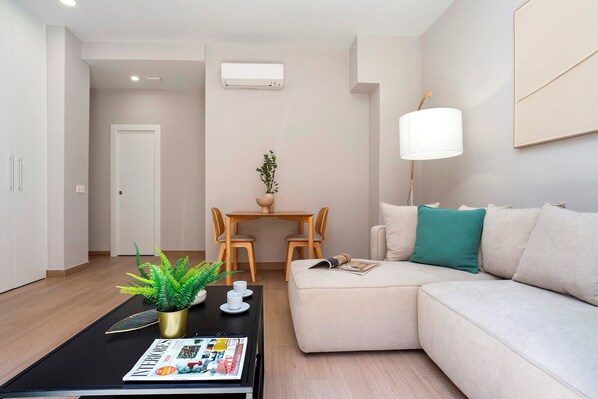 Una sala de estar moderna con un sofá, una mesa de centro con una revista, una planta en maceta y un comedor con mesa y sillas.