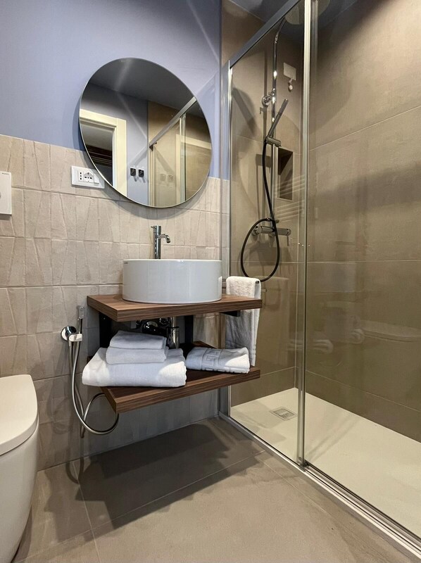Een moderne badkamer met een douchecabine van glas, een ronde spiegel, een wit wastafel en netjes opgevouwen handdoeken op een plank.
