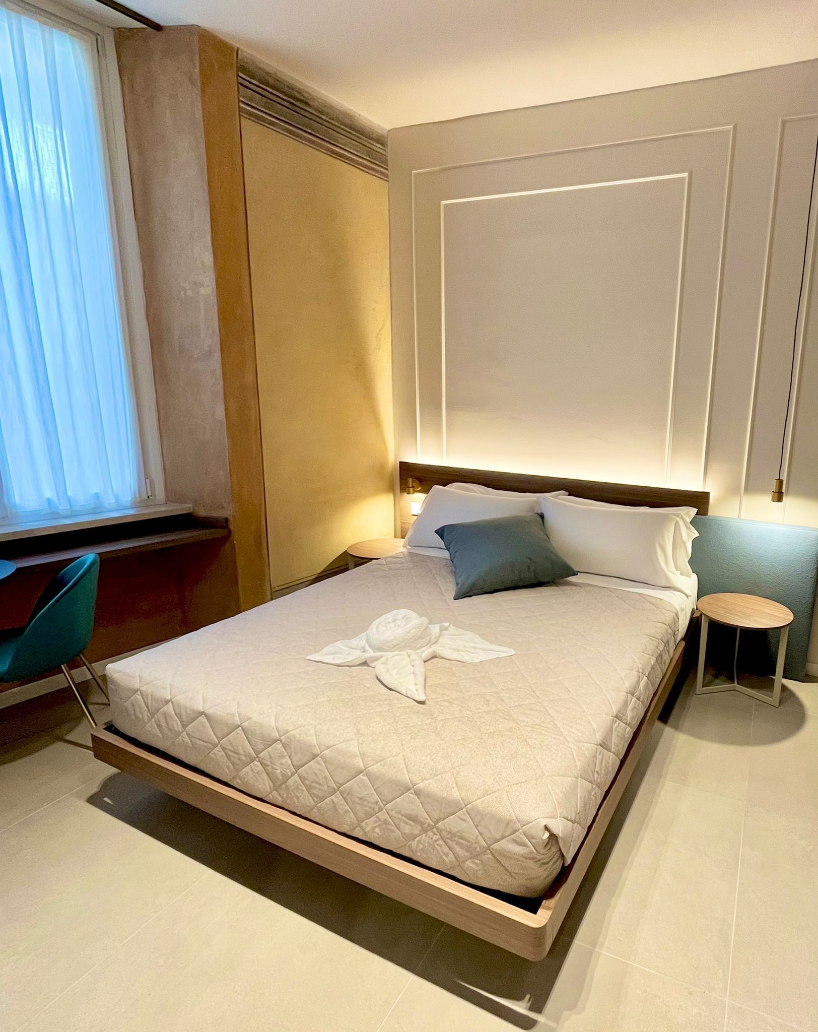 A neatly made bed with a quilted cover, a bedside table, a chair, and a window with curtains.