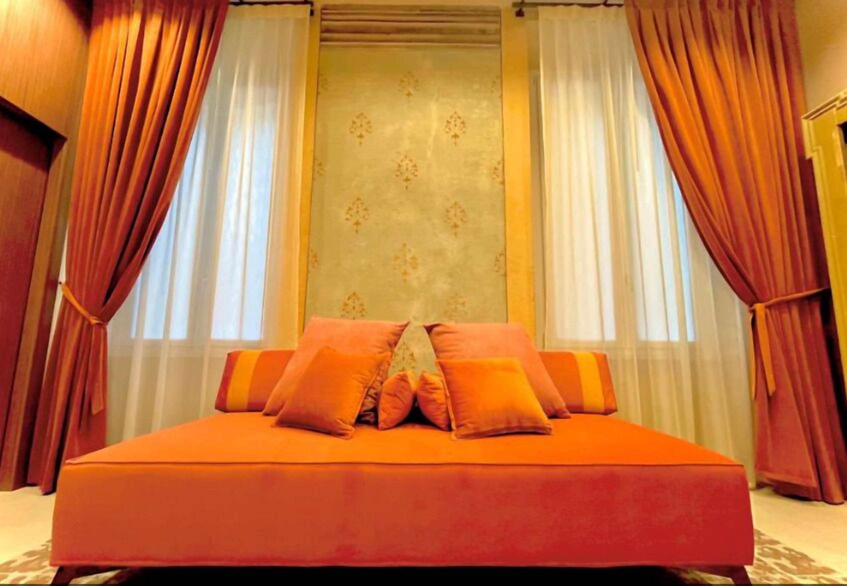 Un letto grande con biancheria arancione e molti cuscini, affiancato da tende rosse e un muro decorato.
