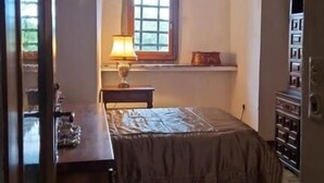 Un dormitorio con piso de madera, una cama con colcha estampada, una mesita de noche, una lámpara, una ventana con vistas y un arco de madera en la entrada.