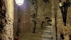 Una escalera de piedra que asciende en un edificio histórico tenuemente iluminado, con un farol y apliques en la pared.