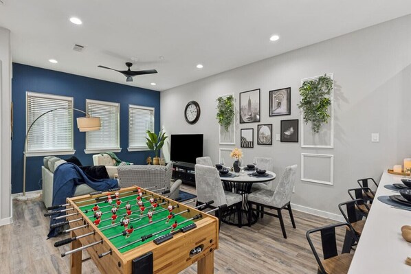 Una sala moderna con mesa de fútbol, un sofá, un comedor con sillas y un televisor.