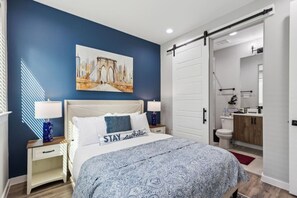 Un dormitorio con una cama, dos mesitas de noche, una mesita auxiliar, un cuadro colgado en la pared, una puerta corrediza hacia el baño y una alfombra roja.