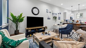 A modern living room with a flat-screen TV, comfortable seating, a coffee table, and a dining area with a kitchen in the background.