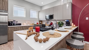 A modern kitchen with a central island, wooden cabinets, and a red accent wall. - The Jasper 2BD TH: 7ST Walkable, Stylish & Homey! (Fort Worth)