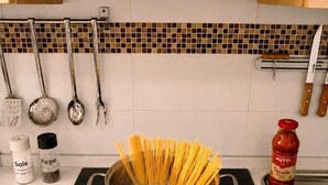 Een keuken met een inductiekookplaat, een pan kokend water, spaghetti, een snijplank met rode chilipepers, een fles Mott's ketchup, en een citroen op het aanrecht.