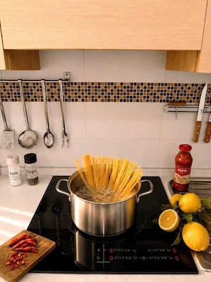 Una cucina con piano cottura a induzione, una pentola di acqua bollente, spaghetti, un tagliere con peperoncini rossi, una bottiglia di ketchup Mott's e un limone sul bancone.