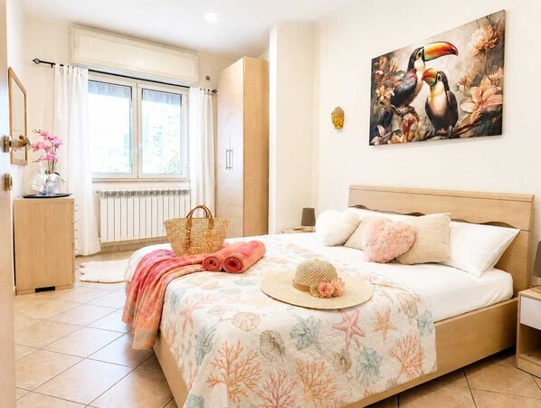 Un dormitorio con cama, ventana, radiador, mesita de noche, lámpara, un cuadro y una lámpara fijada a la pared.