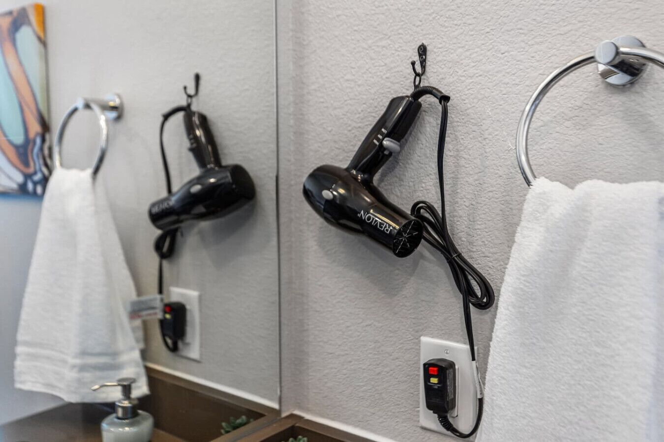 A bathroom with two hair dryers mounted on the wall, a towel rack, and a soap dispenser.