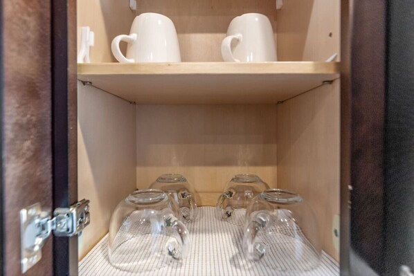 A wooden cabinet with two white mugs on the top shelf and four clear glass cups on the bottom shelf.