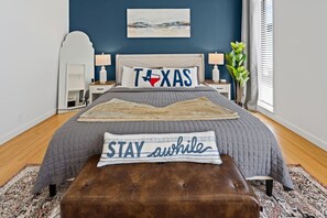 A bedroom with a bed, a nightstand, a lamp, a plant, and a wall with a painting.