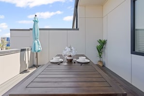 A rooftop terrace with a wooden table set for two, a potted plant, and a light blue umbrella.