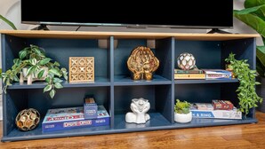 A TV on a stand with a shelf below it containing board games, a decorative globe, and a small plant. - Charming 2BD: TCU Walkable, Blissful & Joyful ! (Fort Worth)