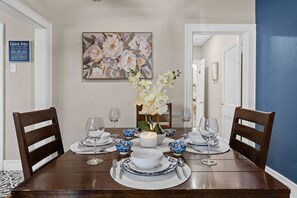 A dining table set with plates, bowls, and wine glasses, surrounded by chairs, with a floral painting and a 'Kitchen Rules' sign on the wall. - This is Your Home Away From Home!  Very Comfy 2BD! (Fort Worth)