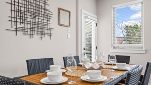 Een eethoek met een houten tafel gedekt voor vier personen, omringd door rieten stoelen. Aan de muur hangt een modern kunstwerk en er is een raam met zonwering.