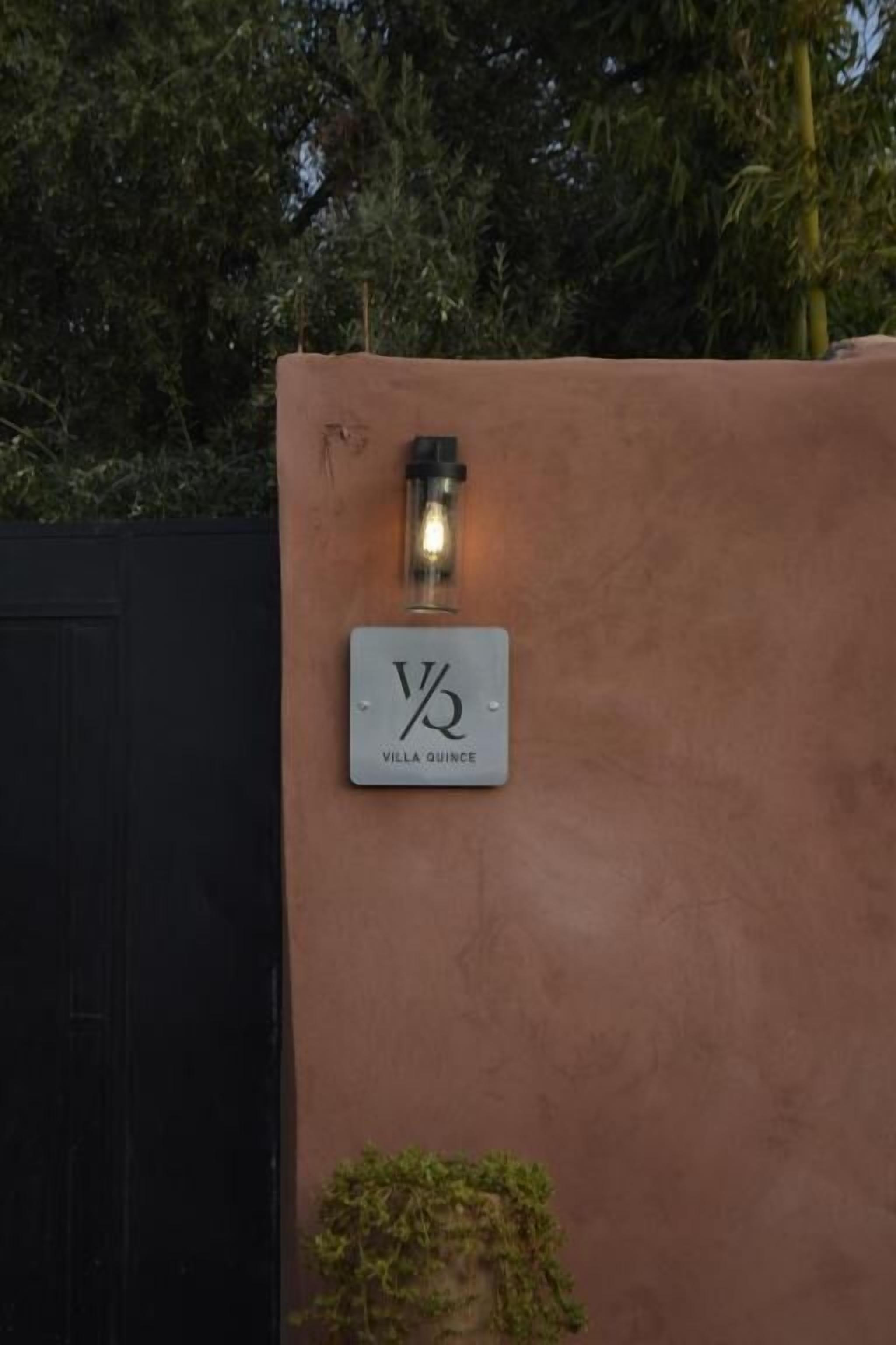 A sign with "VILLA QUENCE" on a textured wall, a light fixture above it, and a small plant below.