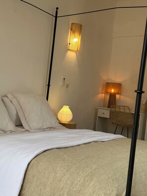 A four-poster bed with white linens, a bedside lamp, a wall-mounted light, and a wicker chair. - Villa Quince  (Ouirgane)