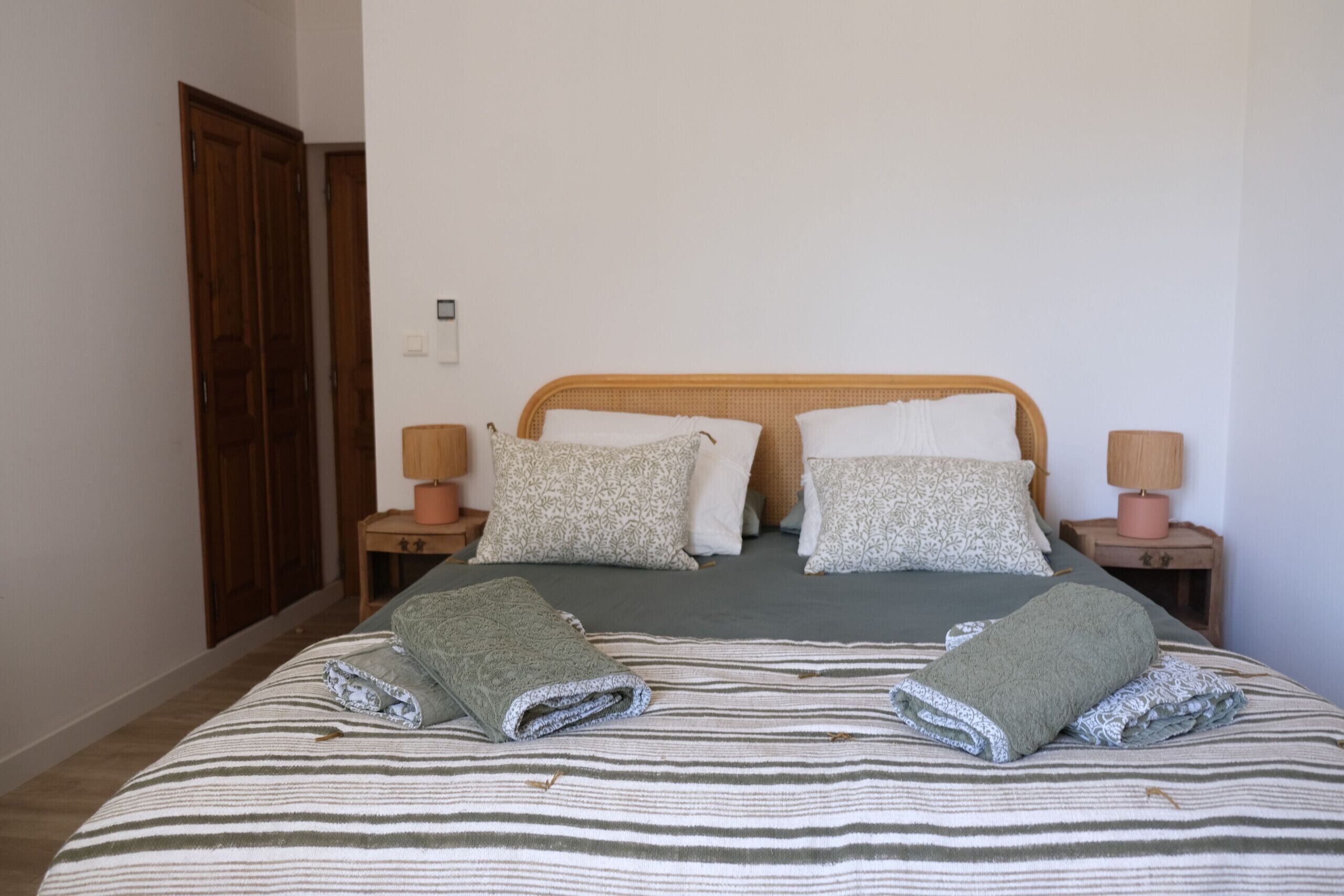 A neatly made bed with striped bedding and two folded towels. There are two bedside tables with lamps, a wooden headboard, and a door in the background.
