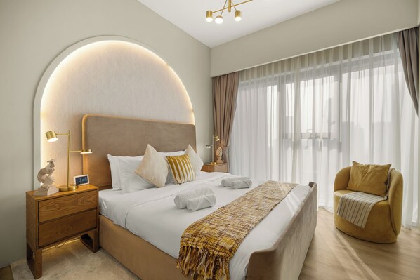 A modern bedroom with a large bed, a nightstand, a chair, and a large window with sheer curtains.