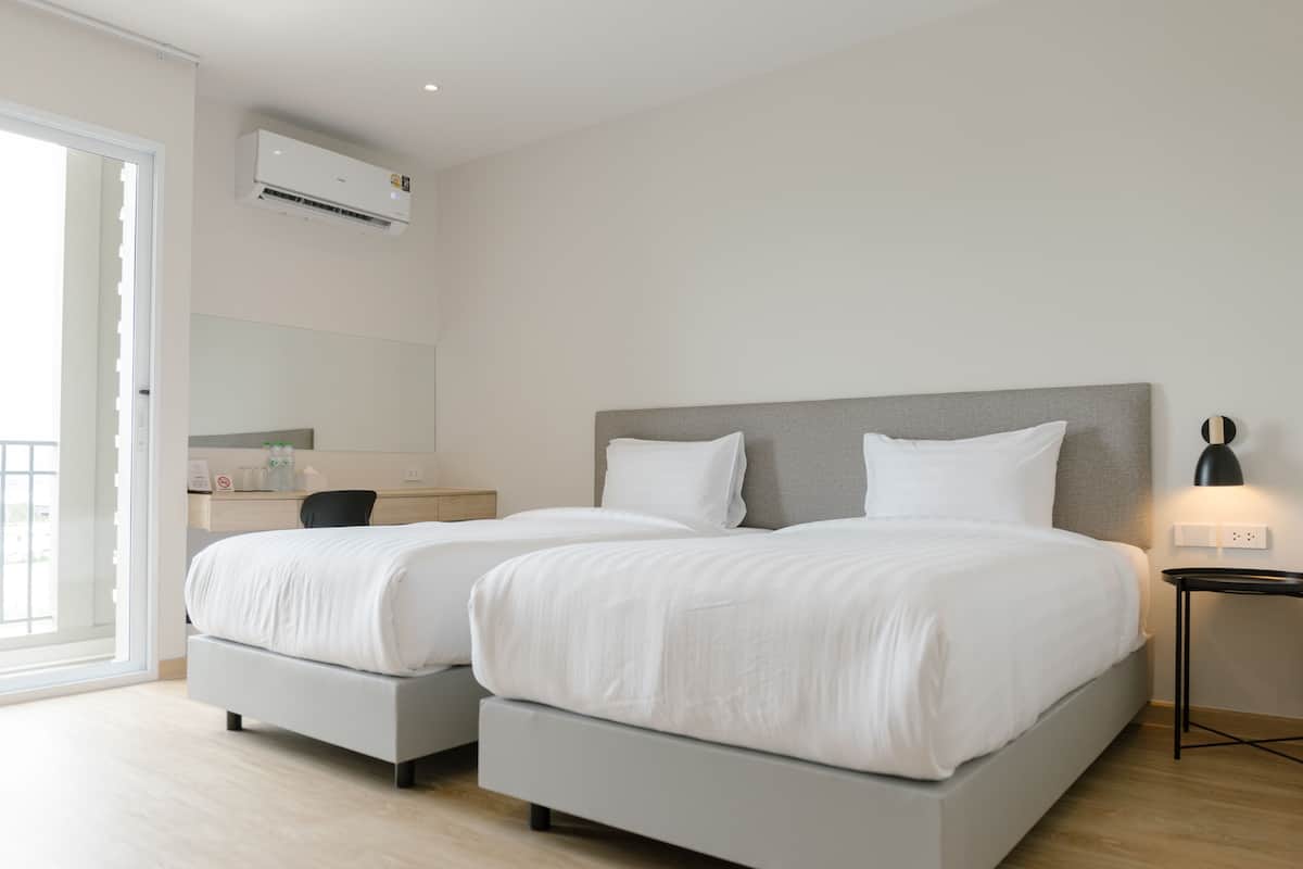 Una habitación de hotel moderna con dos camas, un escritorio, una silla, una mesita de noche, una lámpara y un equipo de aire acondicionado.