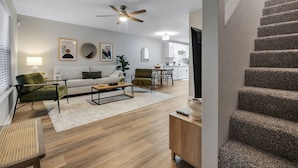 A modern living room with a sofa, armchairs, a coffee table, and a dining area. A staircase leads to an upper floor.