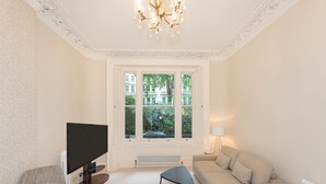 A modern living room with a large window, a flat-screen TV, a sofa, a glass coffee table, and a floor lamp. - Bright and Spacious Studio, Knightsbridge, London - Pass the Keys (London)