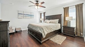 Une chambre à coucher avec un grand lit, un ventilateur de plafond, une table de chevet, une commode et une fenêtre avec des rideaux.