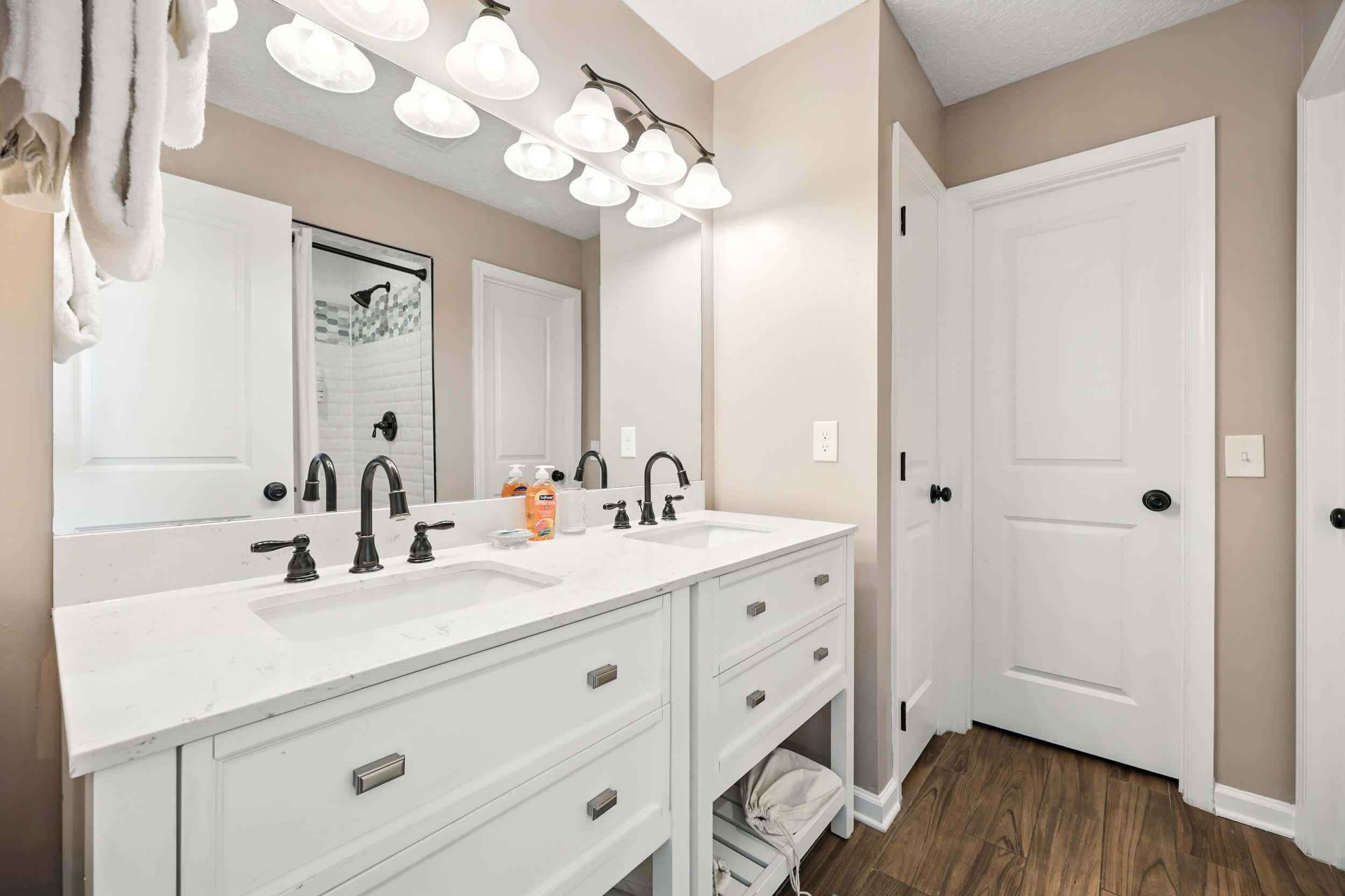 A bathroom with a double sink, white vanity, and a large mirror. There are multiple light fixtures above the mirror and a towel rack to the left.