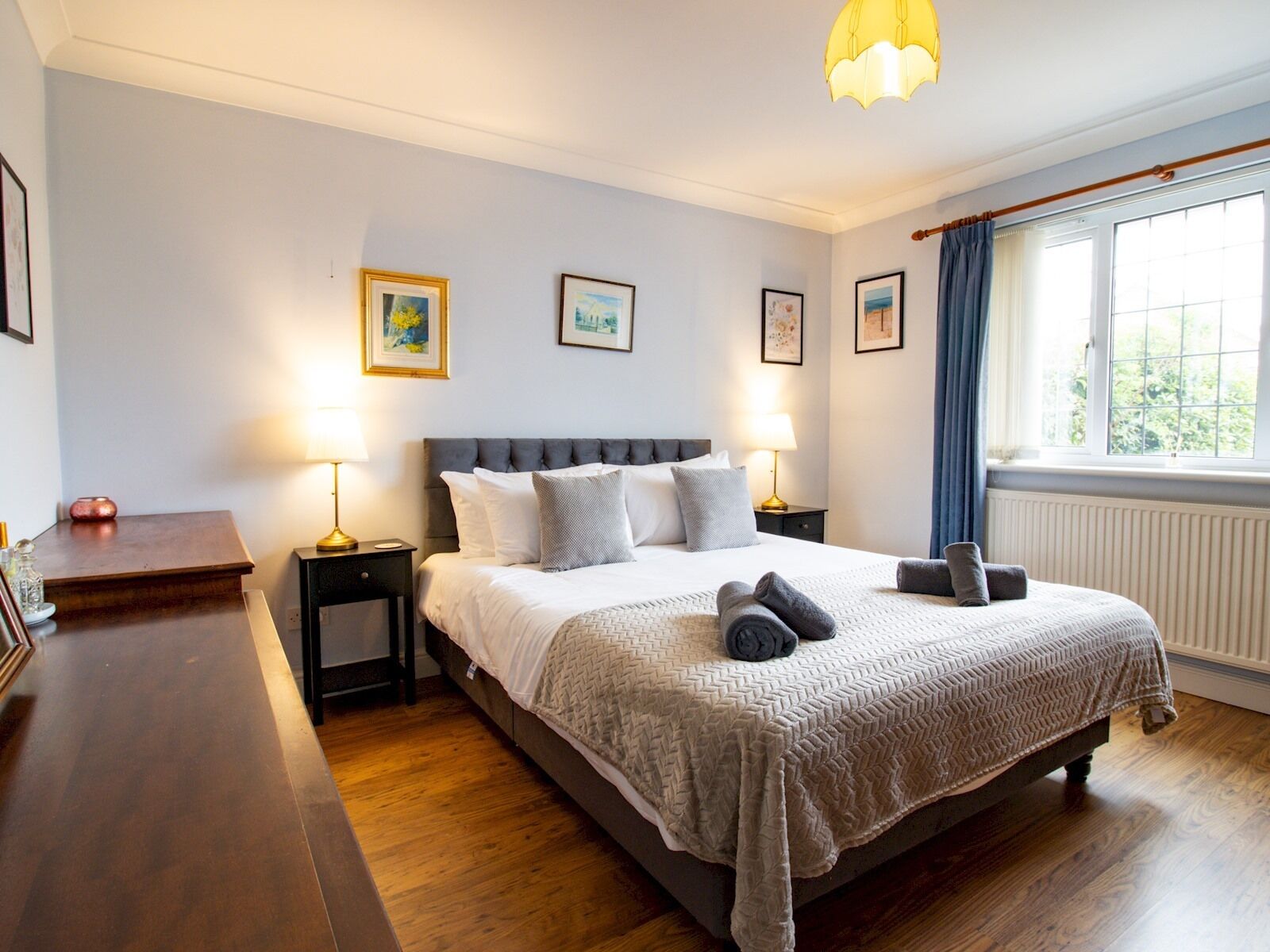 Une chambre à coucher bien rangée, avec un grand lit, des lampes de chevet, des tableaux encadrés au mur et une fenêtre avec des rideaux.