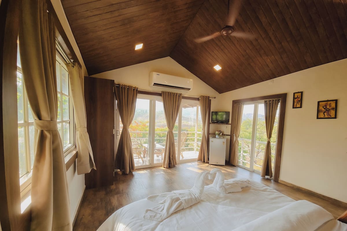 A hotel room with a bed, curtains, a ceiling fan, an air conditioning unit, a television, and a balcony with a view.