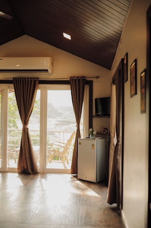 A room with a wooden ceiling, a sliding glass door with curtains, a small refrigerator, a television on a shelf, and a chair outside the door. - Green View Resort (Karjat)