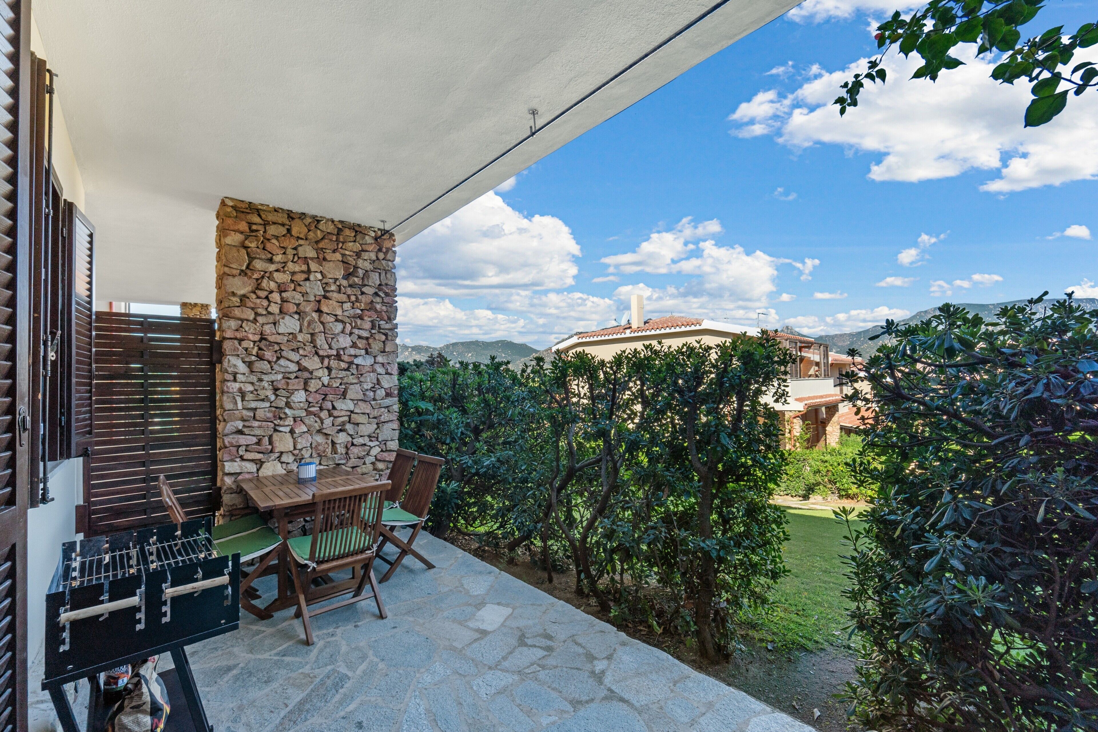Eine Terrasse mit Steinmauer, Tisch und Stühlen, mit Blick auf einen Garten und ein entferntes Gebäude.