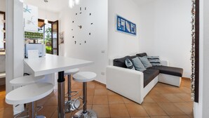 A modern living space with a white sofa, a dining table with bar stools, a wall clock, and a framed artwork.